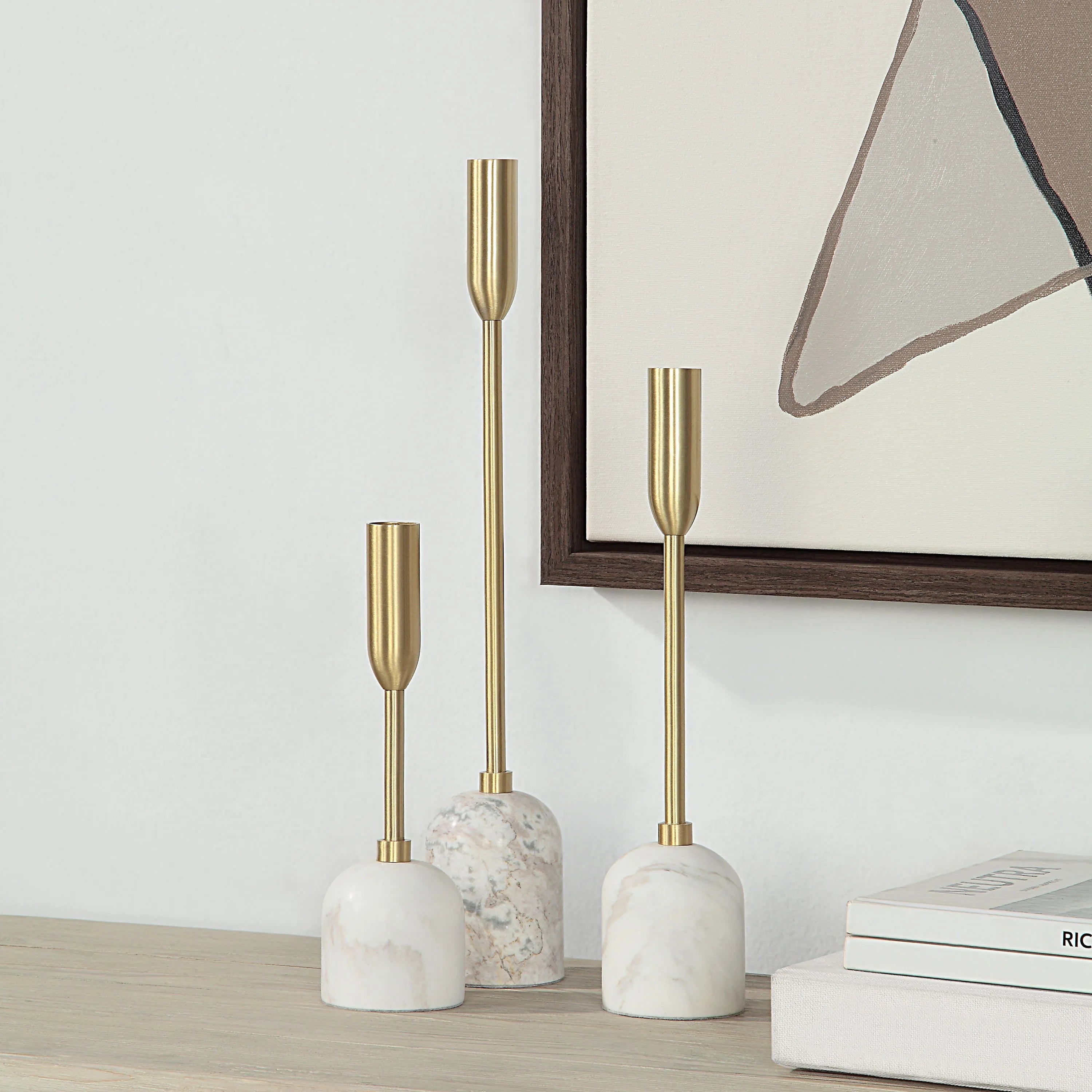 Aurelia Candle Holders with white marble bases and brass stems displayed on a wooden surface beside framed art and books