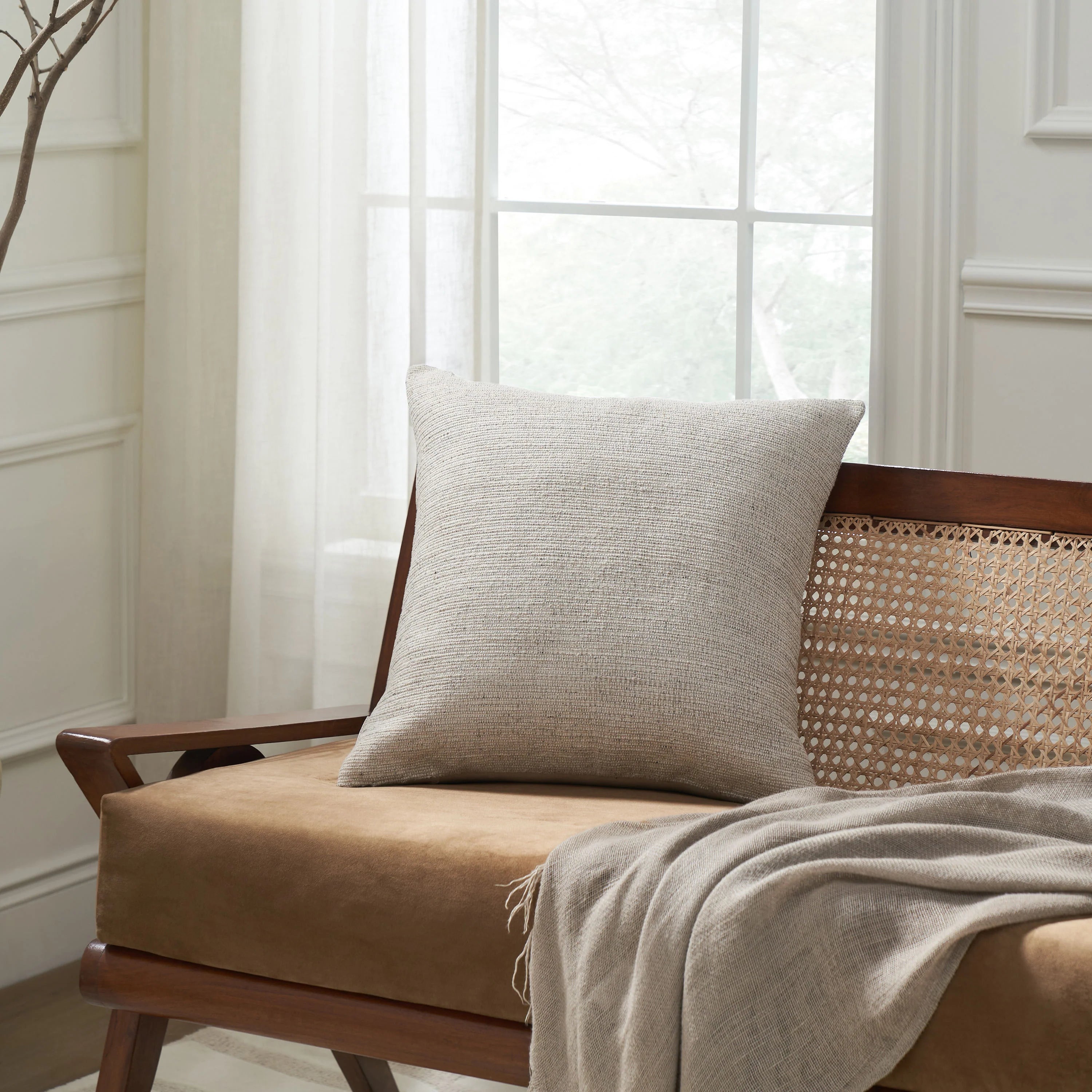 The warm beige Bennett stone mix pillow styled on a wooden cane-back bench with a soft grey throw blanket.