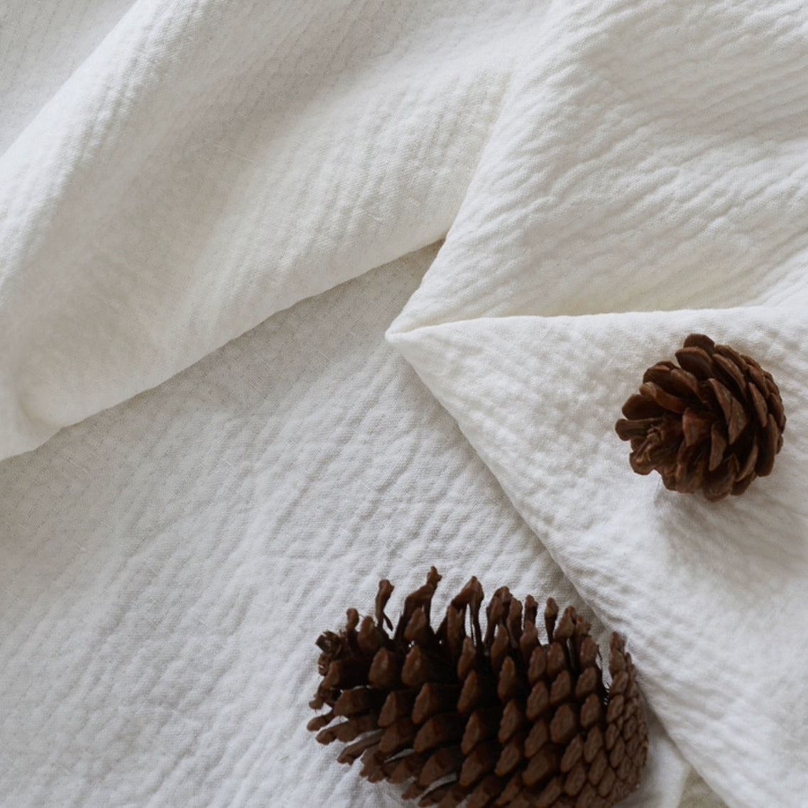 Close-up of breathable cotton muslin fabric in soft white showing natural texture and weave detail with pinecones for styling accent
