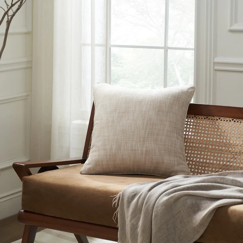 The light beige Eloise woven pillow styled on a warm brown cushion of a wooden cane-back bench near a bright window.