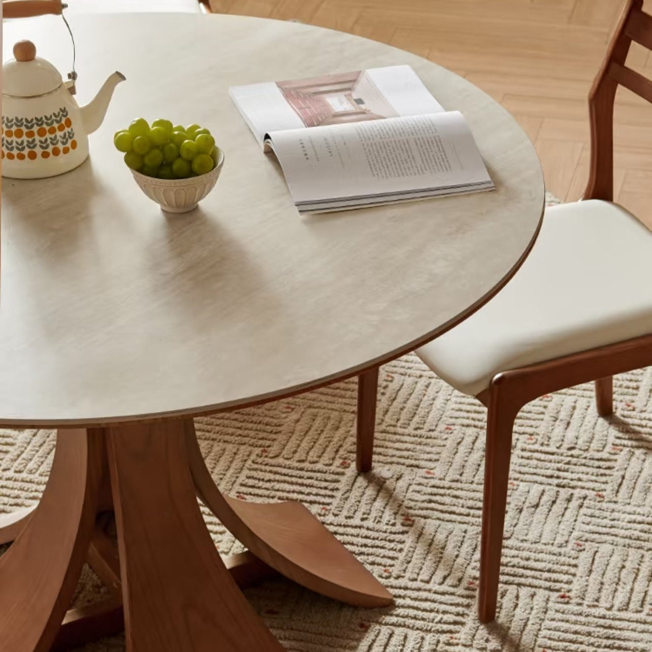 Close-up of Elowen Round Dining Table with walnut base, ceramic teapot, grapes, and open book on tabletop