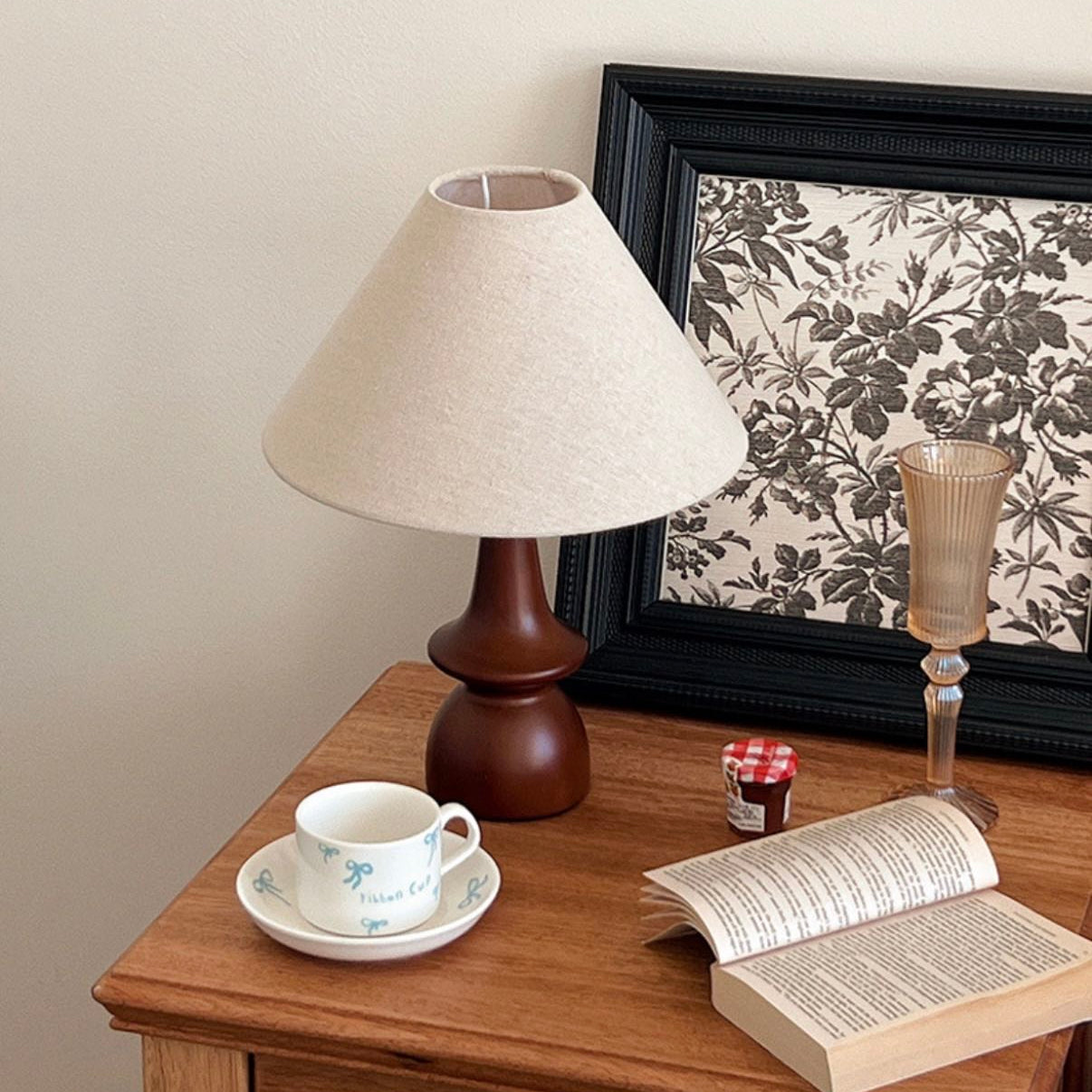 Olivia Table Lamp with wooden base on a bedside table beside a cup, book, and framed floral art