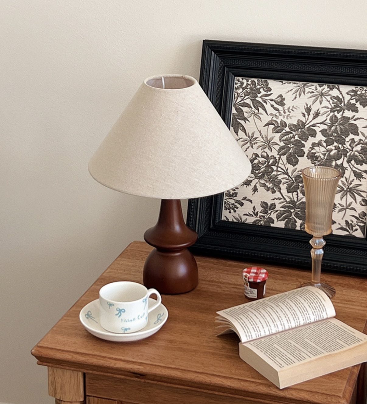Olivia Table Lamp with wooden base on a bedside table beside a cup, book, and framed floral art
