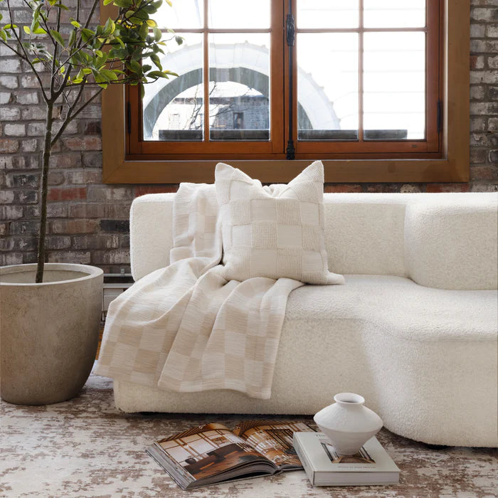 The cream Piper knitted checkerboard pillow styled on a textured white boucle sofa next to a draped throw blanket.