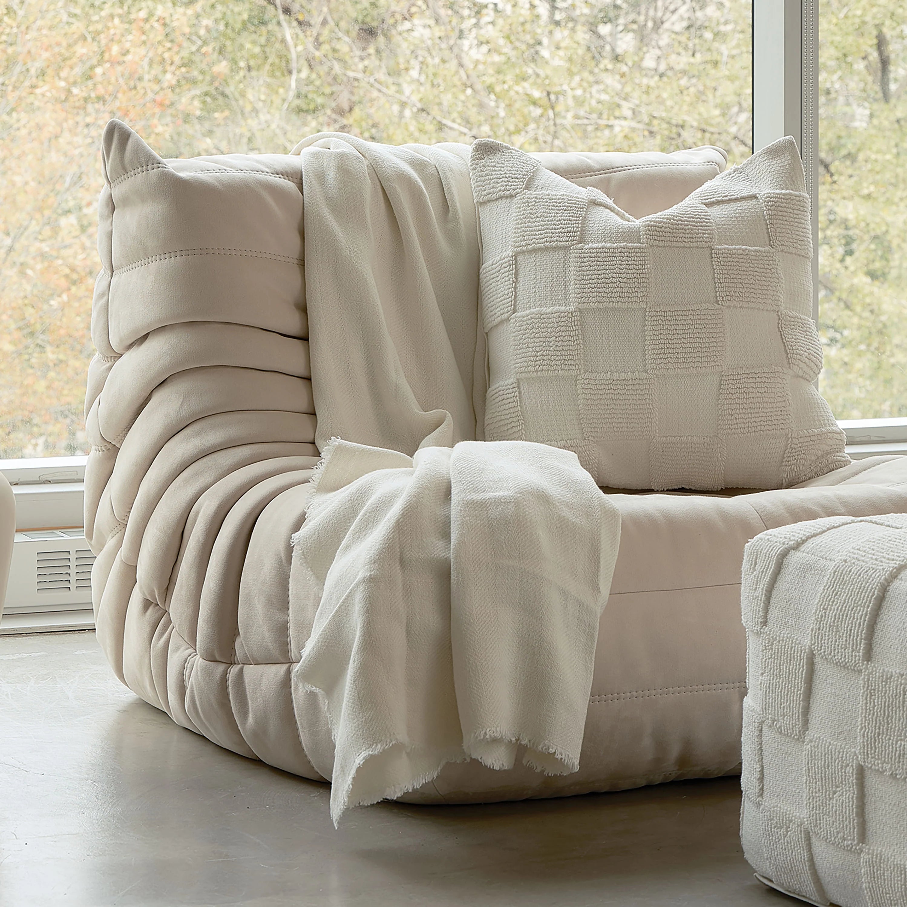 The Piper cream checkerboard throw pillow styled on a modern channeled lounge chair next to a large bright window.