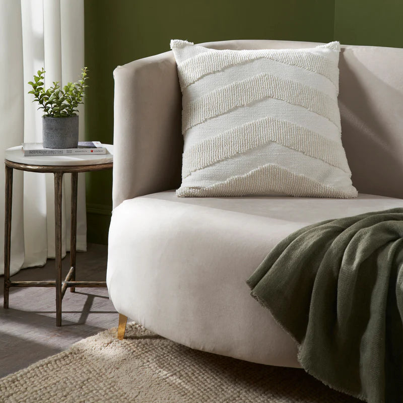 The white Seraphina knitted chevron pillow styled on a beige velvet armchair next to a green throw blanket and olive green wall.
