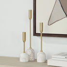 Aurelia Candle Holders with white marble bases and brass stems displayed on a wooden surface beside framed art and books