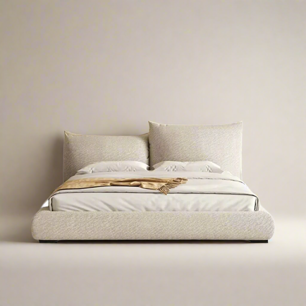 A beige upholstered bed frame with a low-profile design, accompanied by matching pillows and a protective plastic covering on the mattress.