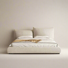 A beige upholstered bed frame with a low-profile design, accompanied by matching pillows and a protective plastic covering on the mattress.