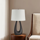Calla Table Lamp with sculpted black wood base and white shade on bedside table beside modern armchair