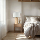 Callisto Travertine Table Lamp on wooden nightstand beside cozy beige bed with soft linens in natural light bedroom