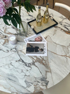 Close-up of Celesti Elegance round dining table with marble-patterned sintered stone top styled with flowers, magazines, and gold bottles