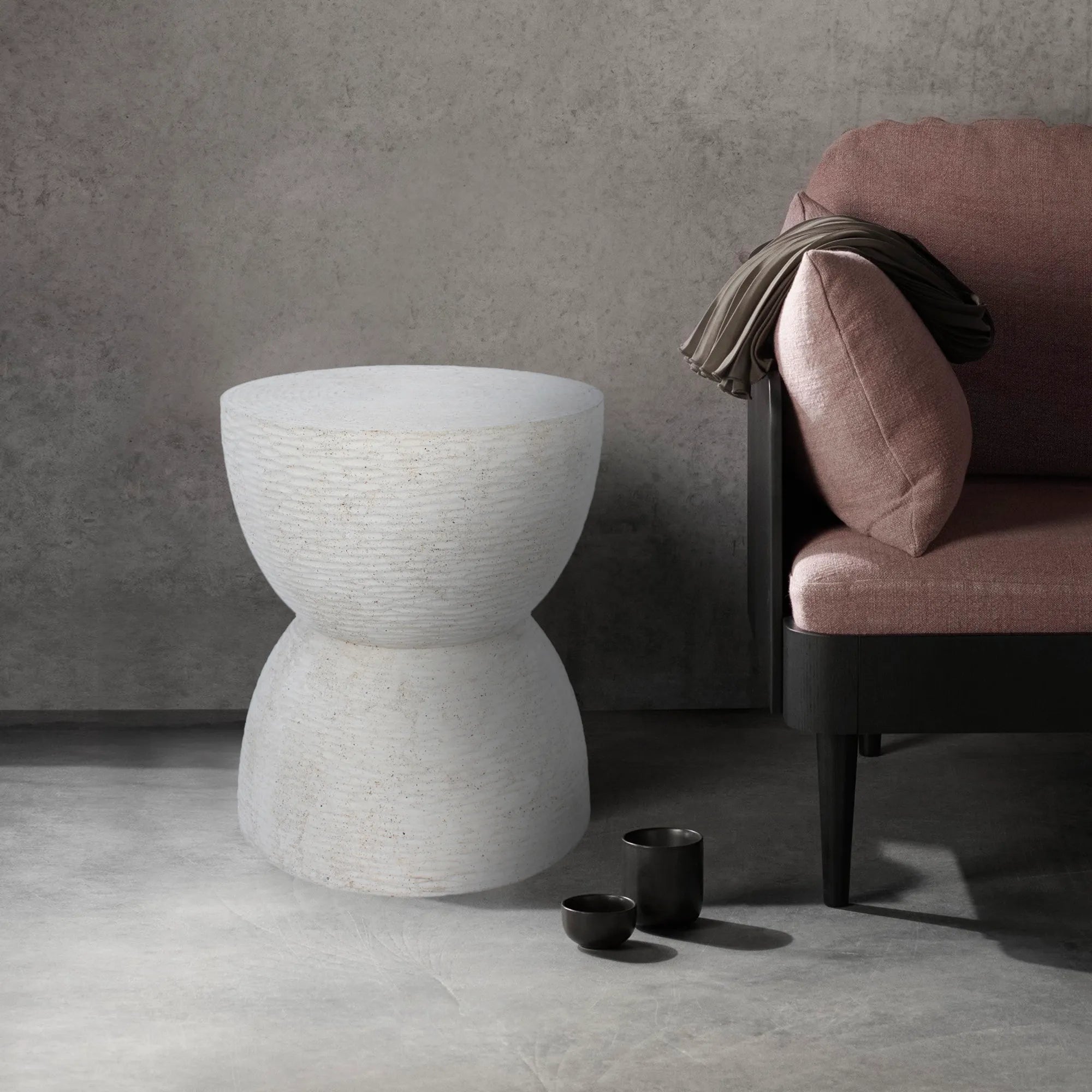 Dune textured stone side table styled next to a mauve armchair in a modern industrial living room with concrete walls.