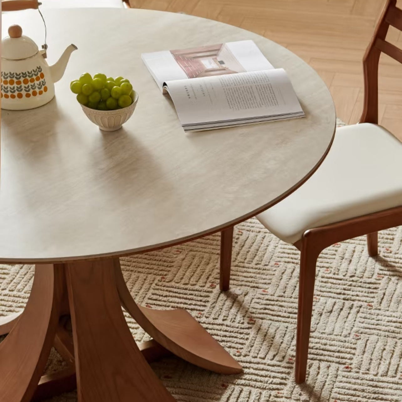 Close-up of Elowen Round Dining Table with walnut base, ceramic teapot, grapes, and open book on tabletop