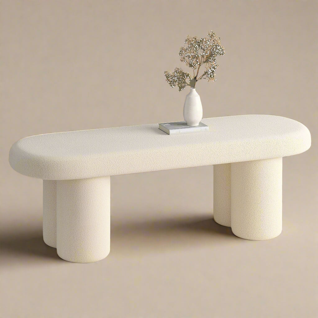 A beige upholstered bench with a bouclé fabric texture, featuring rounded edges and sturdy cylindrical legs, displayed in a room with a decorative vase on top.