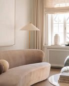 Horizon Shade Floor Lamp with brass stand and wide cone shade placed beside a curved beige velvet sofa in cozy living room