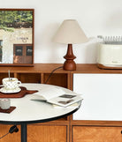 Olivia Table Lamp with walnut base placed on a wooden sideboard beside a table with cup and book