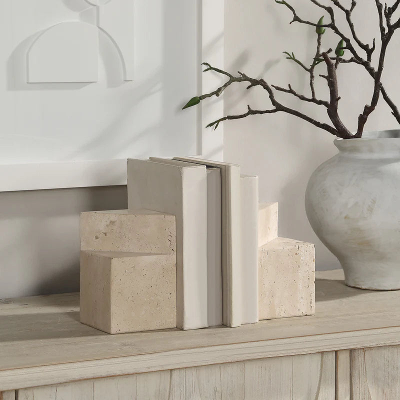 Sculptural geometric travertine bookends styled on a wooden sideboard holding white books, highlighting their functional weight and modern aesthetic.